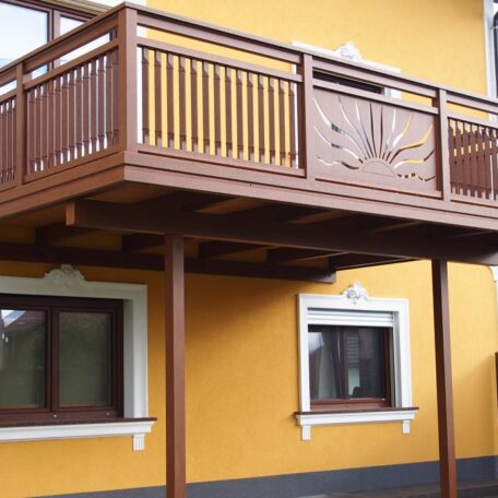 Brauner Holzbalkon mit verziertem Geländer an gelbem Haus, darunter zwei weiße Fenster. HANSEL.kraus metalltechnik gmbh