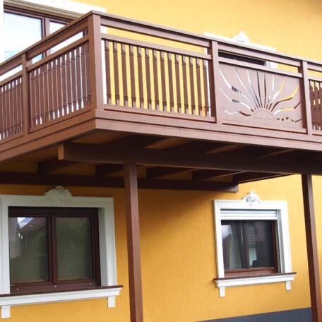 Balkon aus Holz mit detailreichem Geländer an gelber Hausfassade, darunter zwei Fenster. HANSEL.kraus metalltechnik gmbh