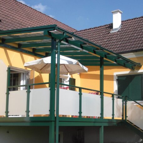 Überdachte Terrasse mit Glasdach, grünem Gestell und Sonnenschirm an gelbem Wohnhaus. de HANSEL.kraus metalltechnik gmbh