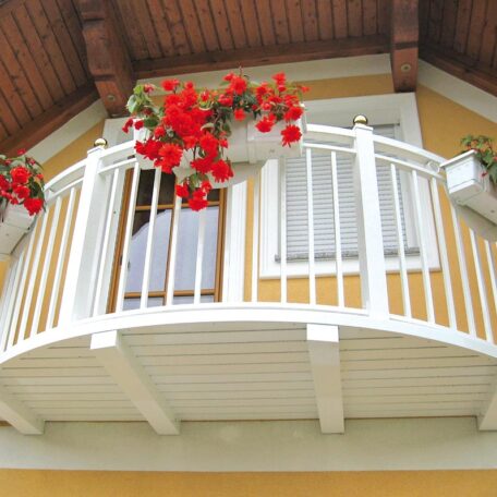 Weißer Balkon mit roten Blumen und Holzüberdachung an gelber Hausfassade. HANSEL.kraus metalltechnik gmbh