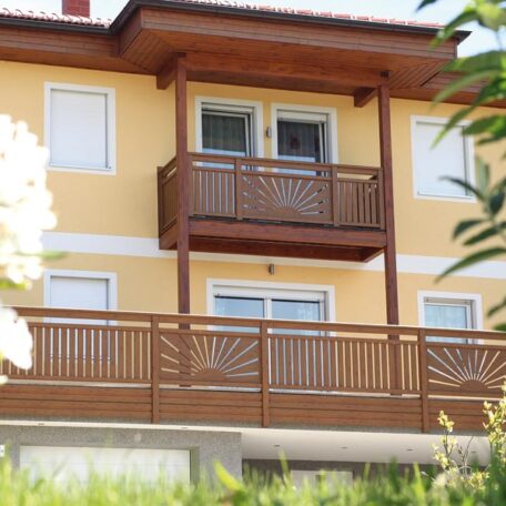 Zweistöckiges gelbes Wohnhaus mit Holzbalkon und geschlossenen Fensterläden, umgeben von grüner Bepflanzung. HANSEL.kraus metalltechnik gmbh