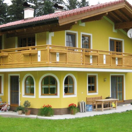 Zweistöckiges gelbes Einfamilienhaus mit Holzbalkon und gepflegtem Garten vor dem Haus. HANSEL.kraus metalltechnik gmbh