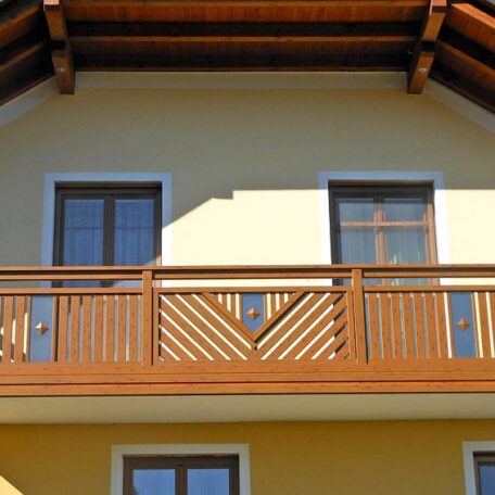Holzbalkon mit Geländer vor zwei Fenstern an gelber Hausfassade unter Satteldach. HANSEL.kraus metalltechnik gmbh
