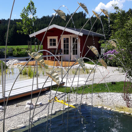 Ein rotes Gartenhaus steht hinter einem Naturteich, umgeben von Pflanzen und Kies. HANSEL.kraus metalltechnik gmbh