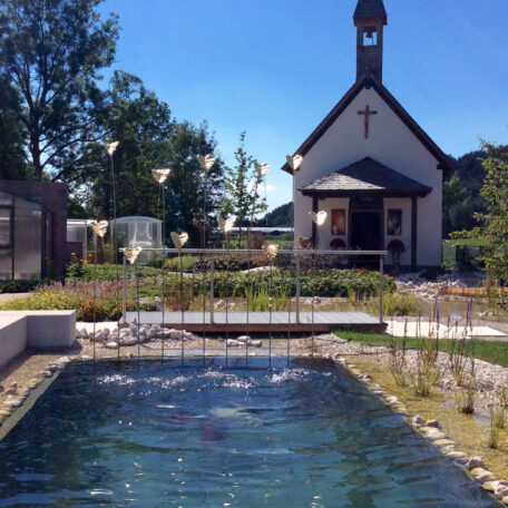 Kapelle mit Kreuz am Teich, umgeben von Gartenanlagen und strahlend blauem Himmel. HANSEL.kraus metalltechnik gmbh