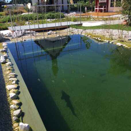 Natürlicher Schwimmteich mit Steinumrandung und Steg im modernen Garten, sonniger Tag. HANSEL.kraus metalltechnik gmbh