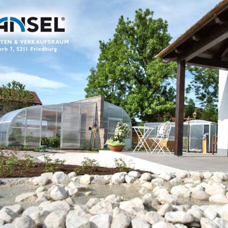 Garten mit modernen Gewächshäusern, Steingarten im Vordergrund, daneben ein Haus und Bäume, sonniges Wetter. HANSEL.kraus metalltechnik gmbh