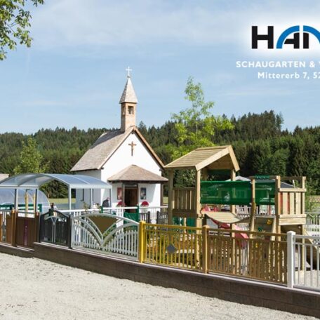 Kinderspielplatz mit Holzkirche, Sandfläche und Klettergerüst bei sonnigem Wetter, im Hintergrund Bäume. HANSEL.kraus metalltechnik gmbh