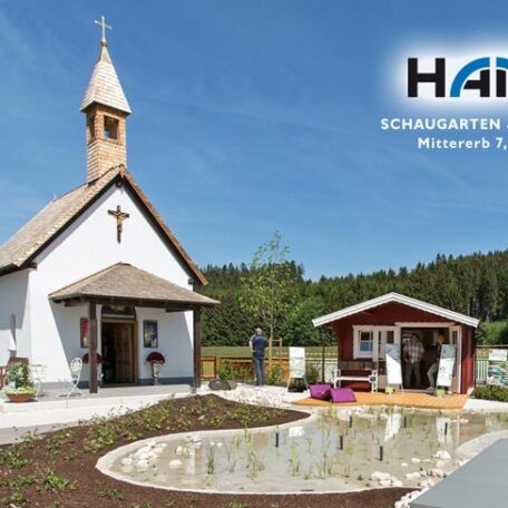 Kapelle mit Garten, kleinem Teich und Infogebäude unter blauem Himmel, umgeben von Wald. HANSEL.kraus metalltechnik gmbh