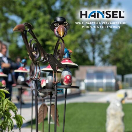 Gartendekorationen aus Metall mit Vogelmotiven stehen auf Rasen, unscharfer Hintergrund mit HANSEL-Logo. HANSEL.kraus metalltechnik gmbh