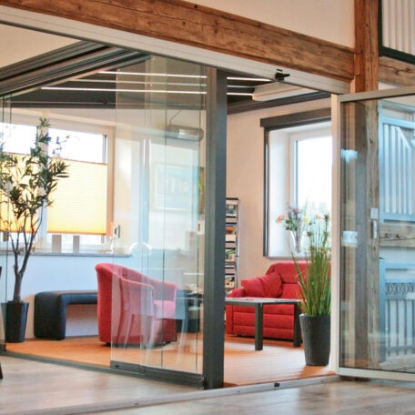 Modernes Büro mit roten Sesseln, Glastür, Holzbalken und großen Fenstern, viel Tageslicht. de HANSEL.kraus metalltechnik gmbh