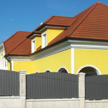 Gelbes Wohnhaus mit rotem Dach, moderner grauer Zaun im Vordergrund, blauer Himmel. HANSEL.kraus metalltechnik gmbh