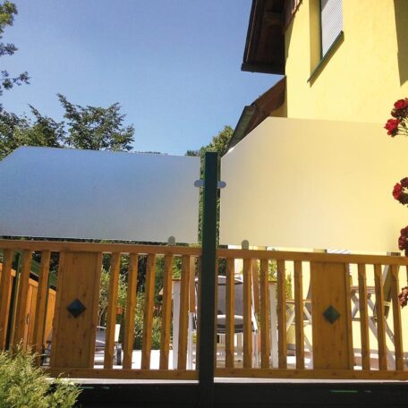 Satiniertes Glasgeländer auf Holzterrasse neben gelbem Haus, Sonnenschein und Blumen am Rand. HANSEL.kraus metalltechnik gmbh