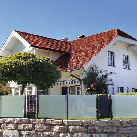 Modernes Einfamilienhaus mit rotem Dach, Garten, Steinmauer und Glaszaun bei sonnigem Wetter. HANSEL.kraus metalltechnik gmbh