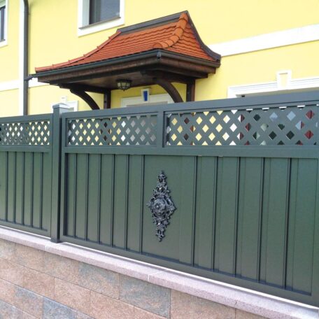 Grüner Metallzaun mit verziertem Element vor gelbem Haus mit Steinsockel und Vordach. HANSEL.kraus metalltechnik gmbh
