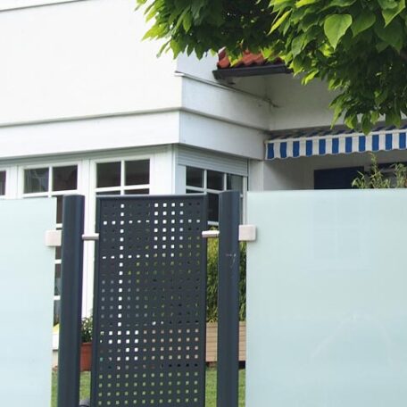 Moderner Metallzaun mit Sichtschutz vor weißem Haus und Markise, grüner Baum im Vordergrund. HANSEL.kraus metalltechnik gmbh