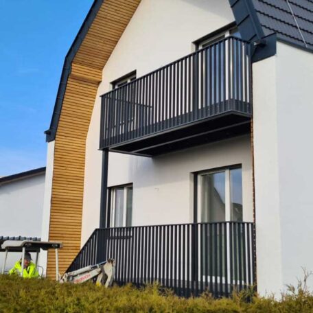 Modernes Wohnhaus mit zwei Balkonen, schwarzen Geländern und Holzfassade an sonnigem Tag. HANSEL.kraus metalltechnik gmbh