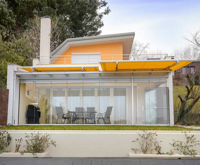 Modernes Einfamilienhaus mit Glas-Terrassenüberdachung und gelber Markise, Garten im Vordergrund. HANSEL.kraus metalltechnik gmbh