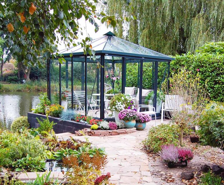 Gartenpavillon mit Glaswänden, umgeben von blühenden Pflanzen und Sitzgelegenheiten am Wasser. de HANSEL.kraus metalltechnik gmbh