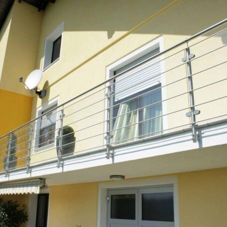 Balkongeländer aus Edelstahl an gelber Hausfassade mit weißem Fenster und Satellitenschüssel. HANSEL.kraus metalltechnik gmbh