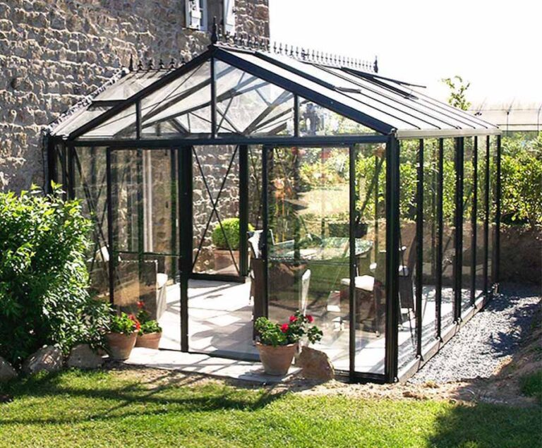 Modernes Glasgewächshaus mit Pflanzen und Sitzbereich im sonnigen Garten neben einem Steingebäude. HANSEL.kraus metalltechnik gmbh