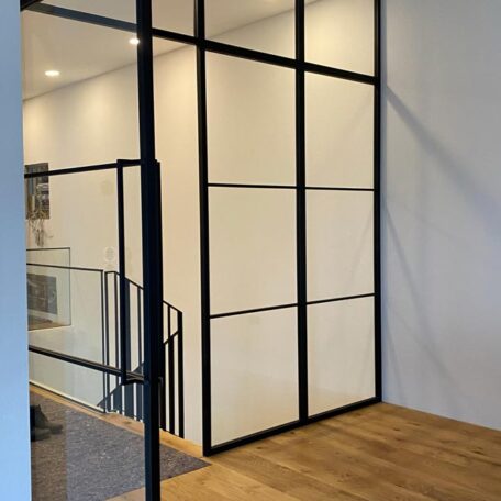 Glaswand mit schwarzem Rahmen trennt modernen Flur vom Treppenhaus, Holzboden sichtbar. HANSEL.kraus metalltechnik gmbh