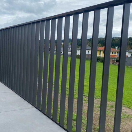 Metallgeländer auf Balkon, daneben grüne Wiese und Häuser im Hintergrund unter bewölktem Himmel. HANSEL.kraus metalltechnik gmbh
