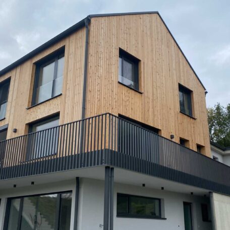 Modernes Haus mit Holzfassade, großen Fenstern und umlaufendem Balkon unter bewölktem Himmel. de HANSEL.kraus metalltechnik gmbh