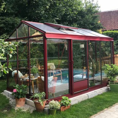 Rotes Gewächshaus im Garten mit Gartenmöbeln und Pflanzen, umgeben von Grün. HANSEL.kraus metalltechnik gmbh