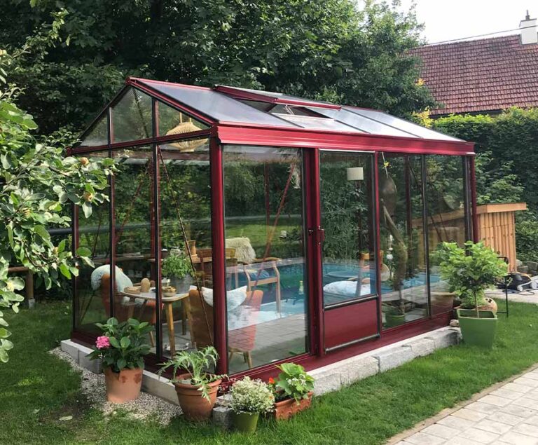 Rotes Gewächshaus im Garten mit Gartenmöbeln und Pflanzen, umgeben von Grün. HANSEL.kraus metalltechnik gmbh