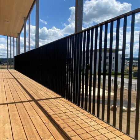 Moderner Balkon mit Holzbelag und schwarzem Geländer, im Hintergrund blauer Himmel mit Wolken. HANSEL.kraus metalltechnik gmbh