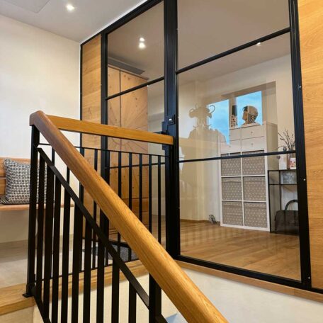Moderne Holztreppe mit schwarzem Geländer vor verglaster Raumtrennung und Regal im Flur. HANSEL.kraus metalltechnik gmbh
