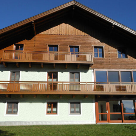 Traditionelles, mehrstöckiges Holzhaus mit Balkon und großen Fenstern bei klarem Himmel. HANSEL.kraus metalltechnik gmbh