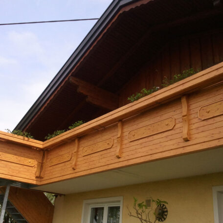 Holzbalkon mit Blumen auf gelbem Haus, traditioneller Stil, blauer Himmel im Hintergrund. HANSEL.kraus metalltechnik gmbh