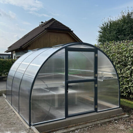 Gewächshaus mit gebogenem Dach aus transparentem Polycarbonat steht im Garten vor einem Wohnhaus. HANSEL.kraus metalltechnik gmbh