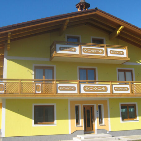 Gelbes Haus im alpenländischen Stil mit Holzbalkonen und weißen Fensterrahmen bei sonnigem Wetter. HANSEL.kraus metalltechnik gmbh