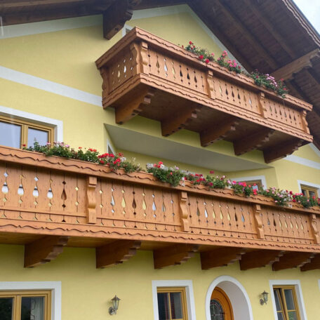 Holzbalkon mit Blumen an gelber Hausfassade, traditionelle Architektur, sonniger Tag. HANSEL.kraus metalltechnik gmbh