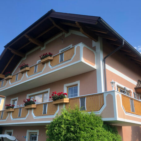 Zweistöckiges Haus mit Holzbalkonen, Blumenkästen und Satteldach vor blauem Himmel. HANSEL.kraus metalltechnik gmbh