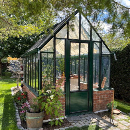Ein Gewächshaus mit Glashauskonstruktion steht im sonnigen Garten neben blühenden Pflanzen. HANSEL.kraus metalltechnik gmbh