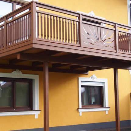 Holzbalkon mit geschnitztem Sonnenmotiv an gelber Hausfassade, darunter zwei Fenster. HANSEL.kraus metalltechnik gmbh