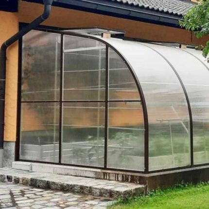Kunststoff-Gewächshaus steht an gelbem Wohnhaus mit Steinweg und grünem Garten. HANSEL.kraus metalltechnik gmbh