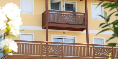 Zweistöckiges, gelbes Haus mit Holzgeländern, Balkon und weißen Fensterläden im Sonnenschein. HANSEL.kraus metalltechnik gmbh