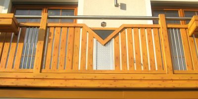 Holzbalkon mit geometrischem Muster vor hellgelber Hauswand und Tür, Tageslicht. HANSEL.kraus metalltechnik gmbh