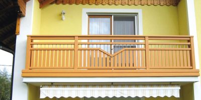 Oberer Holzbalkon mit Geländer vor gelber Hauswand und Glas-Schiebetür, Markise unten. HANSEL.kraus metalltechnik gmbh