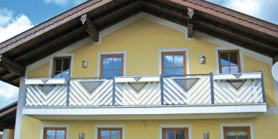 Fassade eines gelben Hauses mit Holzbalkon und Satteldach unter blauem Himmel. HANSEL.kraus metalltechnik gmbh