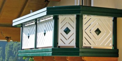Traditionell verzierter Holzbalkon mit grün-weißen Paneelen und geometrischem Muster, sonnig beleuchtet. de HANSEL.kraus metalltechnik gmbh