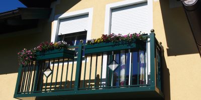 Balkon mit grünem Geländer, bunten Blumen und zwei geschlossenen Rollläden an gelber Hausfassade. HANSEL.kraus metalltechnik gmbh